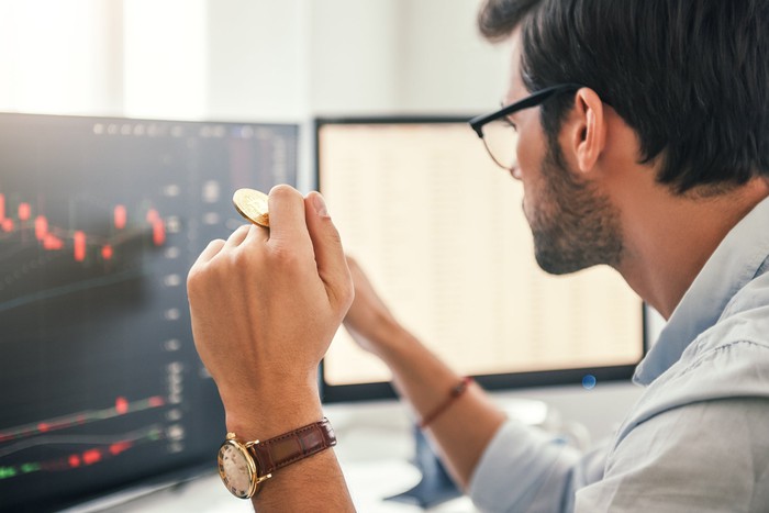 An investor holds a golden coin while looking at a chart on a computer.